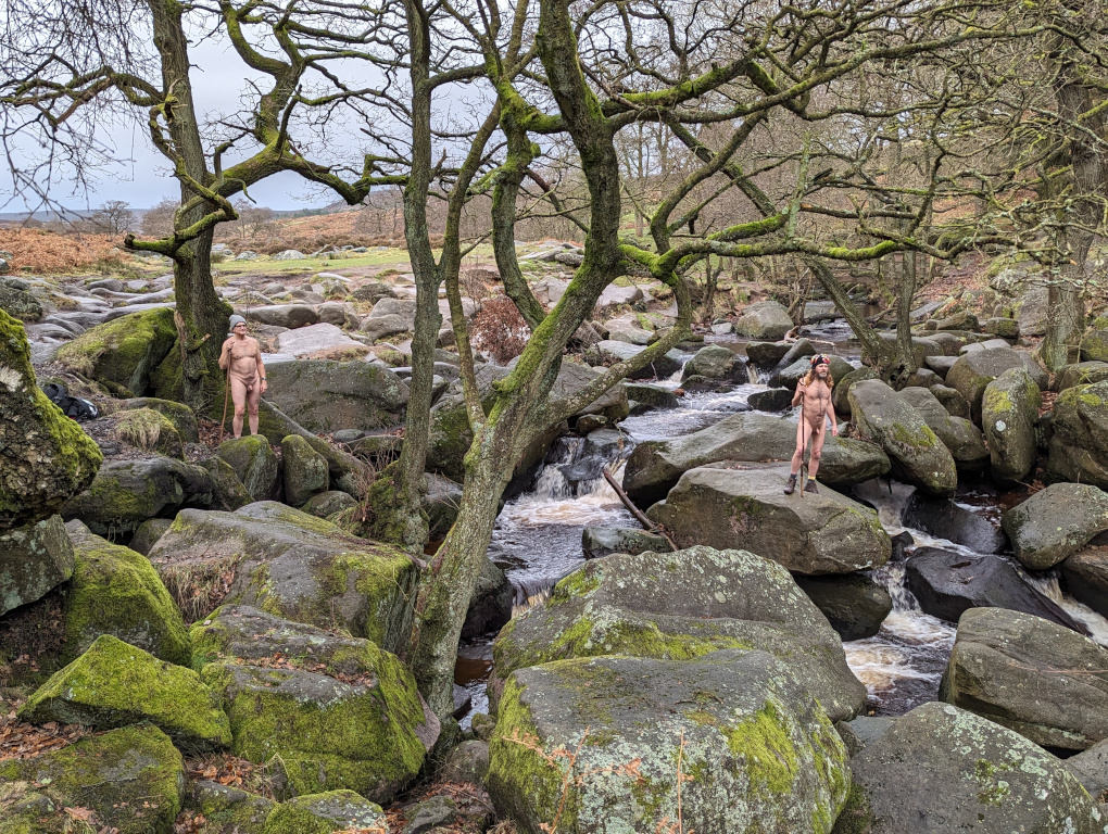 A landscape scene showing a small river running through large boulders, with moss-covered trees bearing no leaves. There are two male figures standing some distance apart on the boulders wearing only hats and footwear and holding long wooden staffs in their hands.